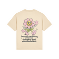 Back view of a cream-colored t-shirt featuring a large retro cartoon pink flower character with a hula hoop, sparkles, and the text “Currently oscillating between delusion and manifestation” in bold black lettering.